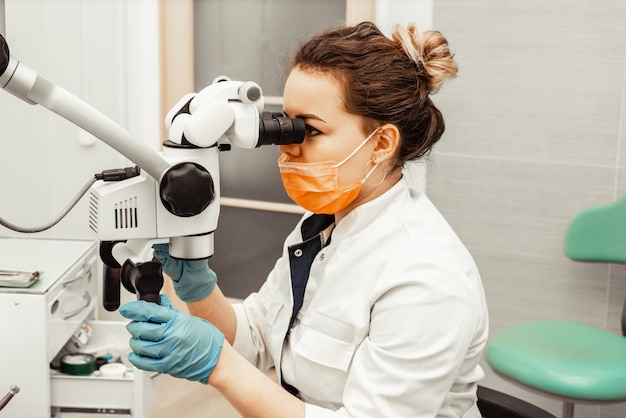 Médico dentista jovem olha através de um microscópio profissional em uma clínica odontológica. Um médico em uma máscara médica descartável e boné. Equipamento avançado em odontologia