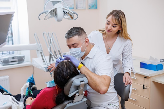 El médico dentista mira los dientes del paciente y sostiene instrumentos dentales cerca de la boca El asistente ayuda al médico Visten uniformes blancos con máscaras y guantes Dentista Consultorio dental