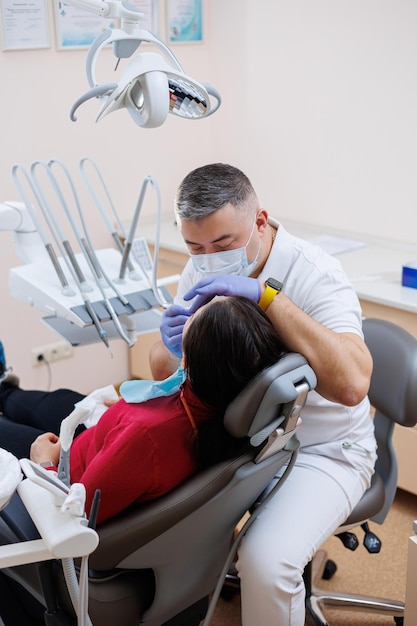 El médico dentista mira los dientes del paciente y sostiene instrumentos dentales cerca de la boca El asistente ayuda al médico Visten uniformes blancos con máscaras y guantes Dentista Consultorio dental