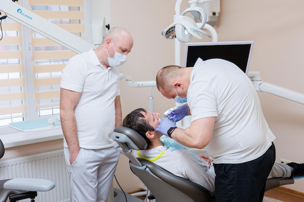 El médico dentista mira los dientes del paciente y sostiene instrumentos dentales cerca de la boca El asistente ayuda al médico Visten uniformes blancos con máscaras y guantes Dentista Consultorio dental