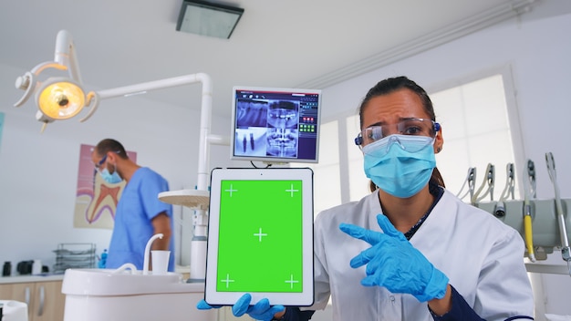 Médico dentista mostrando tableta con pantalla verde, explicando la radiografía dental y el diagnóstico de infección dental. Especialista en estomatología con mascarilla apuntando a maqueta, espacio de copia, pantalla de croma