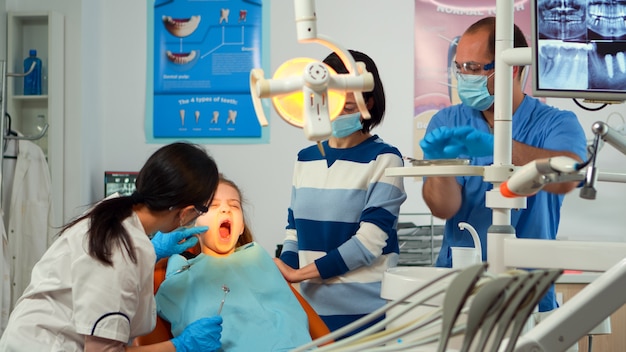 Médico dentista pediátrico que trabaja en la unidad dental con una enfermera y un paciente niña. Estomatólogo hablando con la madre de la niña con dolor de muelas sentada en la silla estomatológica mientras el hombre prepara las herramientas.