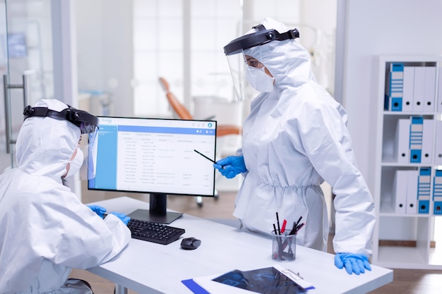 Médico dentista vestindo terno ppe falando com a enfermeira na recepção sobre o calendário dos pacientes. Equipe médica usando equipamento de proteção contra pandemia de coronavírus na recepção odontológica como medida de segurança.