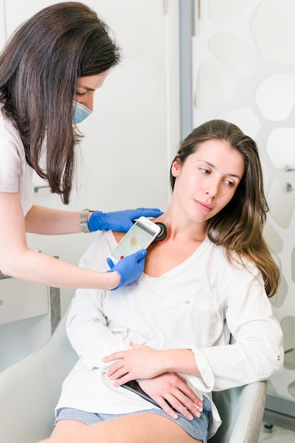 El médico dermatólogo examina la piel de la mujer y el lunar con un aparato especial.