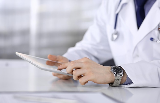 Médico desconhecido sentado e trabalhando com computador tablet na clínica em seu local de trabalho, close-up. Jovem médico no trabalho. Serviço médico perfeito, conceito de medicina.