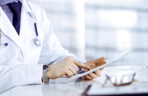 Médico desconhecido sentado e trabalhando com computador tablet na clínica em seu local de trabalho, close-up. Jovem médico no trabalho. Serviço médico perfeito, conceito de medicina.