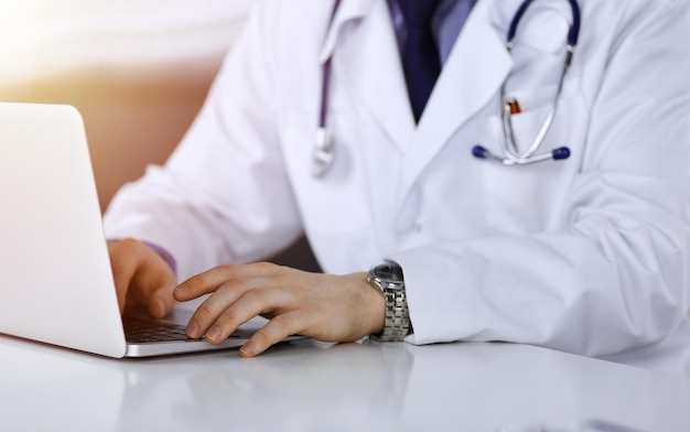 Médico desconhecido sentado e trabalhando com laptop em uma clínica escura, brilho de luz no fundo, close-up de mãos.