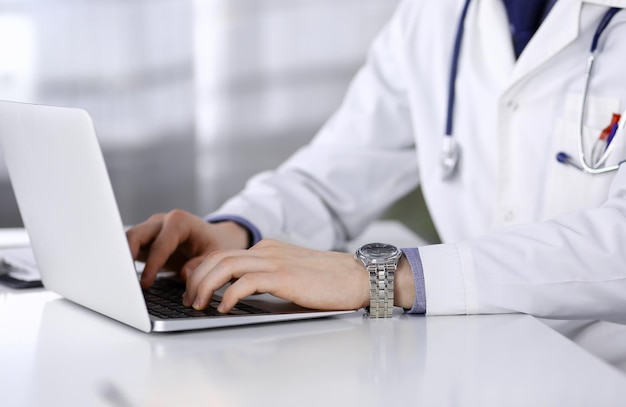 Médico desconhecido sentado e trabalhando com laptop na clínica em seu local de trabalho, close-up. Jovem médico no trabalho. Serviço médico perfeito, conceito de medicina.