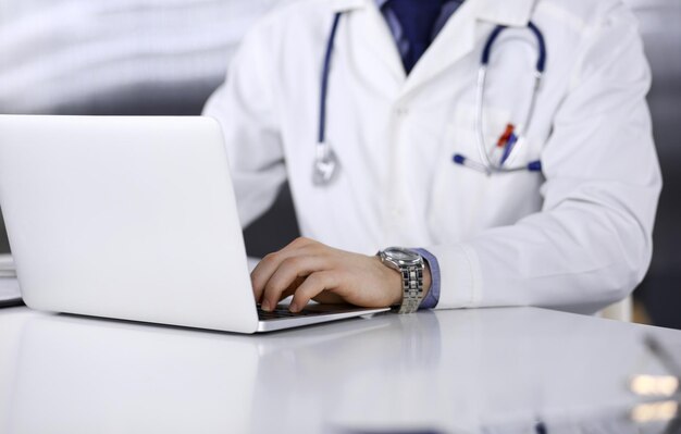 Médico desconhecido sentado e trabalhando com laptop na clínica em seu local de trabalho, close-up. Jovem médico no trabalho. Serviço médico perfeito, conceito de medicina.