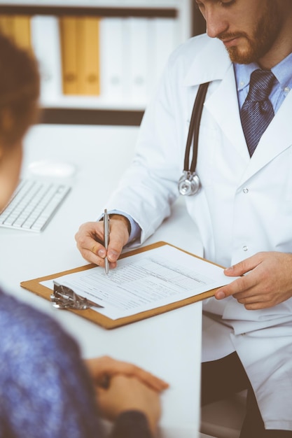 Médico desconocido de barba roja y mujer paciente discutiendo el examen de salud actual mientras está sentado en una clínica soleada, de cerca. concepto de medicina.