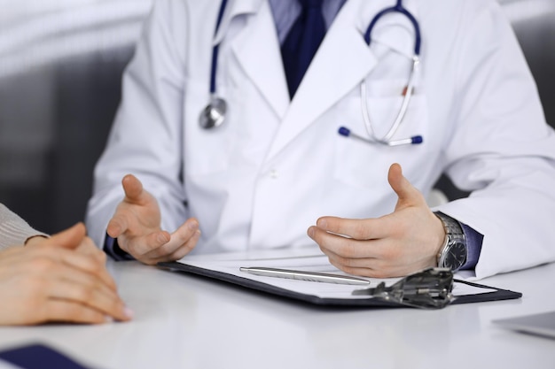 Un médico desconocido y una mujer paciente discutiendo algo mientras estaban sentados en la clínica. El mejor servicio médico en hospital, medicina, parada pandémica.