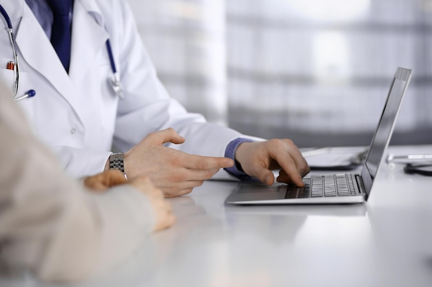 Un médico desconocido y una mujer paciente discutiendo algo mientras estaban sentados en la clínica y usando una laptop. El mejor servicio médico en hospital, medicina, parada pandémica.