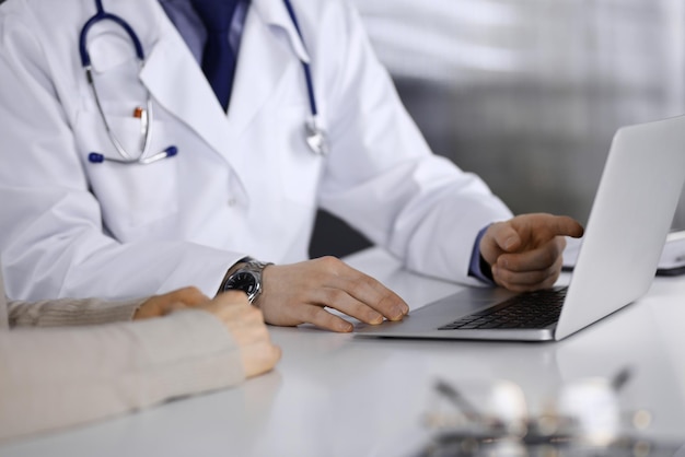 Un médico desconocido y una mujer paciente discutiendo algo mientras estaban sentados en la clínica y usando una laptop. El mejor servicio médico en hospital, medicina, parada pandémica.