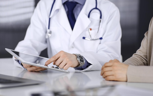 Un médico desconocido y una mujer paciente discutiendo algo mientras estaban sentados en la clínica y usando una tableta. El mejor servicio médico en hospital, medicina, parada pandémica.