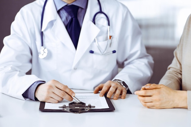 Un médico desconocido y una mujer paciente discutiendo algo mientras se sientan en la clínica y usan el portapapeles. El mejor servicio médico en hospital, medicina, parada pandémica.