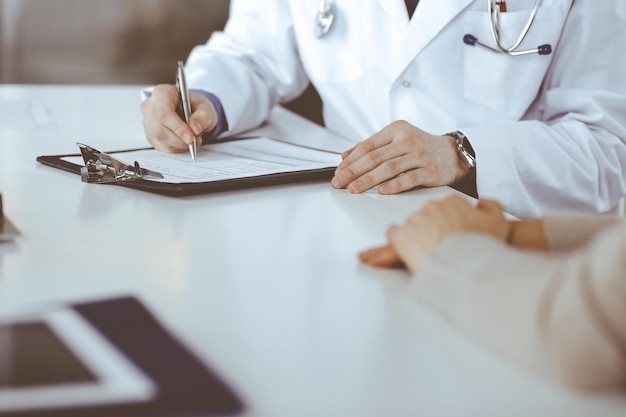 Un médico desconocido y una mujer paciente discutiendo algo mientras se sientan en la clínica y usan el portapapeles. El mejor servicio médico en hospital, medicina, parada pandémica.