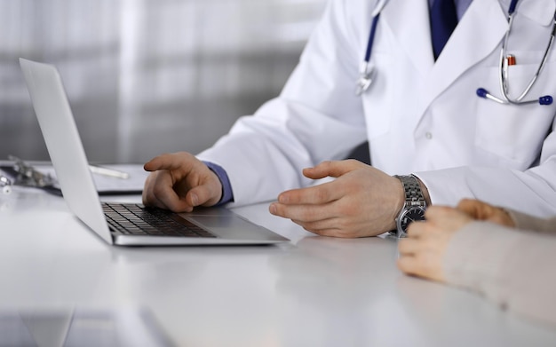 Un médico desconocido y una paciente discutiendo algo mientras están sentados en la clínica y usando una computadora portátil. El mejor servicio médico en hospital, medicina, parada pandémica.