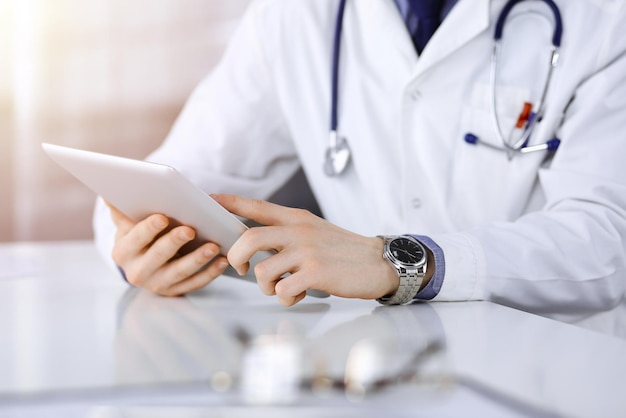 Médico desconocido sentado y trabajando con una tableta en una clínica oscura, deslumbramiento de luz en el fondo, primer plano de manos