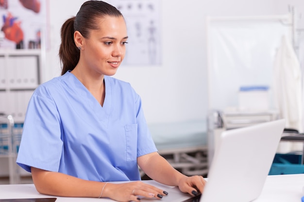 Médico digitando no laptop sentado na mesa usando uniforme azul. Médico de saúde usando o computador na clínica moderna, olhando para o monitor, medicina, profissão, esfrega.