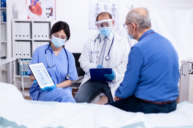 Médico dizendo a um idoso que ele está sofrendo de osteoporose durante o exame médico em um quarto de hospital usando máscara protetora contra covid-19