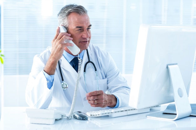 Médico do sexo masculino segurando telefone enquanto olha para o computador no hospital