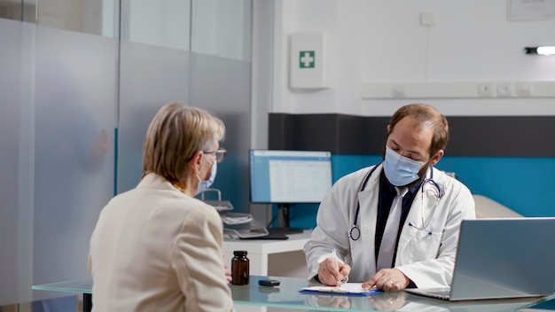 Médico do sexo masculino tomando notas em papéis de prescrição para ajudar o paciente com doença, fazendo exame médico com mulher aposentada. Médico dando tratamento de saúde para idosos com máscara facial.