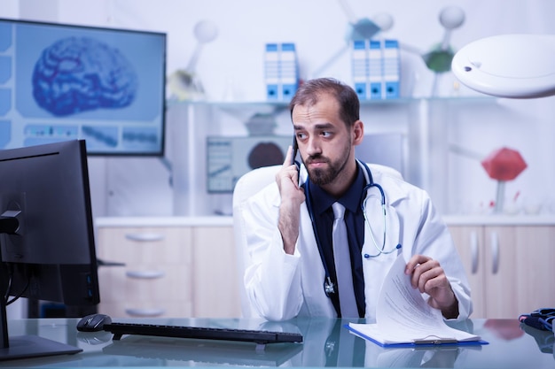 Médico do sexo masculino usando telefone enquanto olha para o monitor do gabinete do hospital. Médico verificando as notas de sua prancheta.