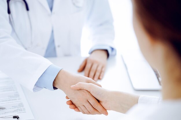 Médico e paciente apertando as mãos enquanto estão sentados um em frente ao outro na mesa da clínica, apenas com as mãos fechadas. Conceito de medicina.