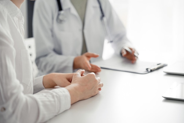 Médico e paciente discutindo o exame de saúde atual enquanto está sentado à mesa no consultório da clínica. O foco está nas mãos da paciente, close-up. Serviço médico perfeito e conceito de medicina