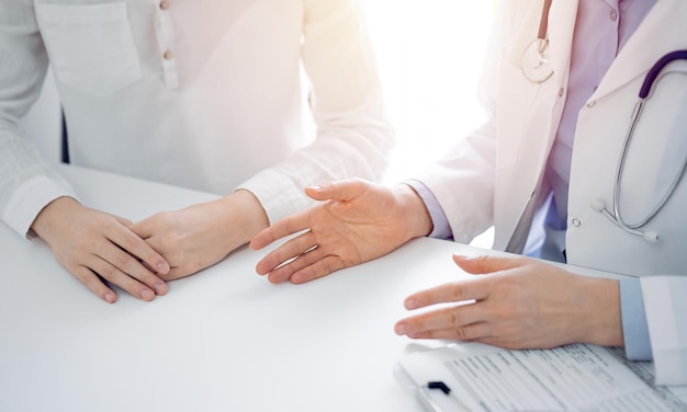 Médico e paciente discutindo questões atuais de saúde enquanto estão sentados um ao lado do outro na mesa da clínica, apenas mãos fechadas. Conceito de medicina.