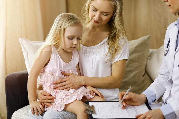 Médico e paciente na sala ensolarada. Pediatra usando a área de transferência enquanto examina a garotinha com a mãe em casa. Conceito de medicina.