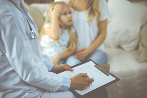 Médico e paciente. Pediatra usando a área de transferência enquanto examina a garotinha com a mãe em casa. Criança doente e infeliz no exame médico. Conceito de medicina