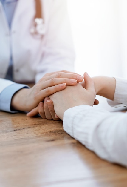 Médico e paciente sentado à mesa de madeira na clínica. Mulher tranquilizadora das mãos do médico feminino. Conceito de medicina.