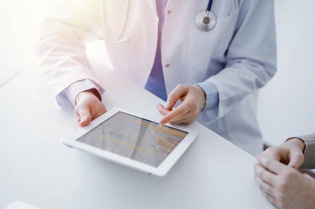 Médico e paciente sentado à mesa na clínica. O foco está nas mãos da médica usando computador tablet, close-up. Conceito de medicina e saúde