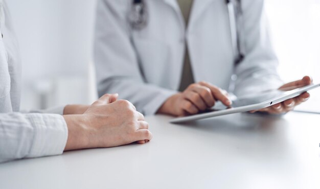 Médico e paciente sentado à mesa no consultório da clínica. O foco está nas mãos da médica usando computador tablet, close-up. Serviço médico perfeito e conceito de medicina