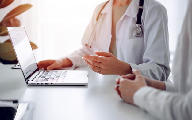 Médico e paciente sentado à mesa no consultório da clínica. O foco está nas mãos da médica usando laptop, close-up. Serviço médico perfeito e conceito de medicina.