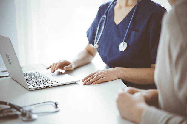 Médico e paciente sentado à mesa no consultório da clínica. O foco está nas mãos de uma médica desconhecida usando um laptop, close-up. conceito de medicina