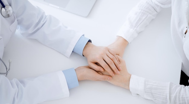 Médico e paciente sentados em frente um do outro à mesa no consultório da clínica. O foco está nas mãos da médica, tranquilizando a mulher, apenas as mãos, de perto. conceito de medicina