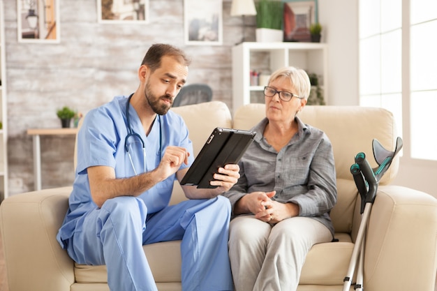 Médico em lar de idosos usando computador tablet enquanto toma com a aposentada.