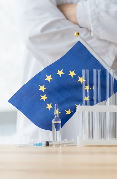 Foto el médico se encuentra cerca del tubo de ensayo, la ampolla, la jeringa y la bandera de la unión europea, el concepto de vacunación global.