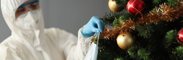 Médico de enfermedades infecciosas en traje protector y mascarilla respiratoria colgante en el árbol de Navidad