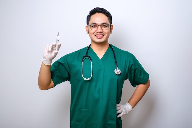 Médico enfermero asiático confiado con batas verdes y guantes de goma sosteniendo una jeringa con la vacuna de pie manteniendo la mano en la cadera