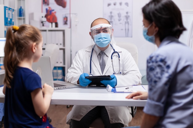 Médico escrevendo diagnóstico de criança em tablet conversando com mãe para tratamento iniciado