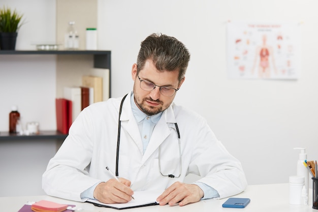 Médico escrevendo em seu caderno no gabinete médico, médico barbudo em uniforme médico branco e óculos sentado no gabinete médico com estetoscópio e trabalhando