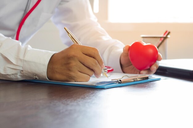 Médico escrevendo receita em papel e segurando um coração vermelho na mesa de trabalho