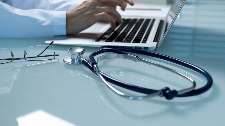 Un médico escribiendo en una computadora portátil con un estetoscopio sobre la mesa.