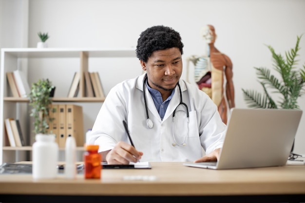 Médico escribiendo en la computadora portátil y tomando notas en la clínica