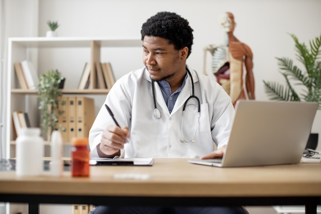 Médico escribiendo en la computadora portátil y tomando notas en la clínica