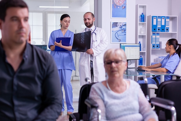 Foto médico especialista que comunica el examen de rayos x a la enfermera mientras escribe el tratamiento en el portapapeles contra la enfermedad para una mujer mayor en silla de ruedas