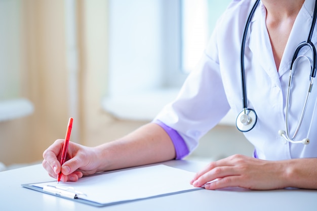 Médico con un estetoscopio escribiendo quejas de pacientes, diagnóstico y tratamiento de la enfermedad en un portapapeles durante una consulta médica.